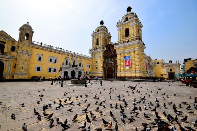 Yellow facade and twin towers of the San Francisco church and convent in Lima, with pigeons on the square