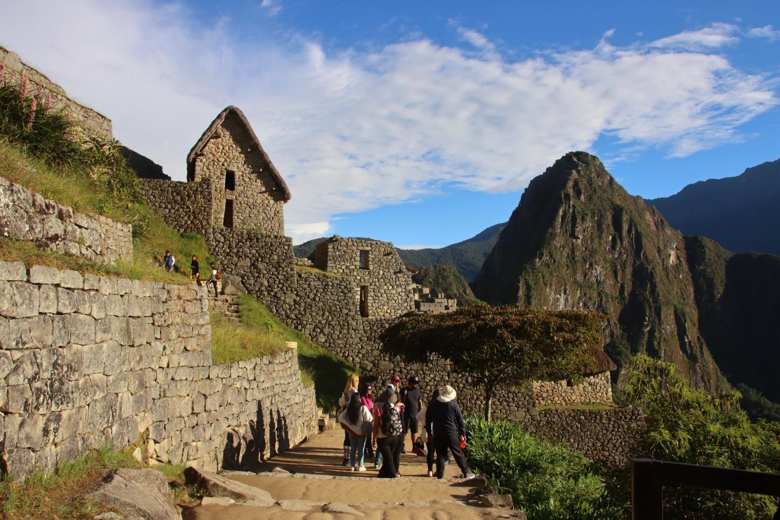 Visitors walking up a stone path toward the guardhouse at Machu Picchu, with Huayna Picchu behind