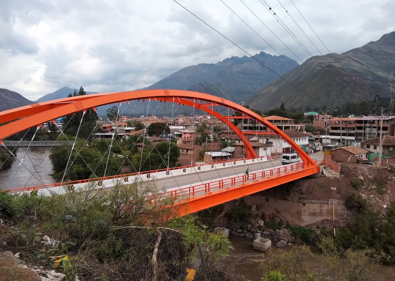 Orange arch bridge over the Urubamba River leading into the town of Urubamba, with green mountains behind