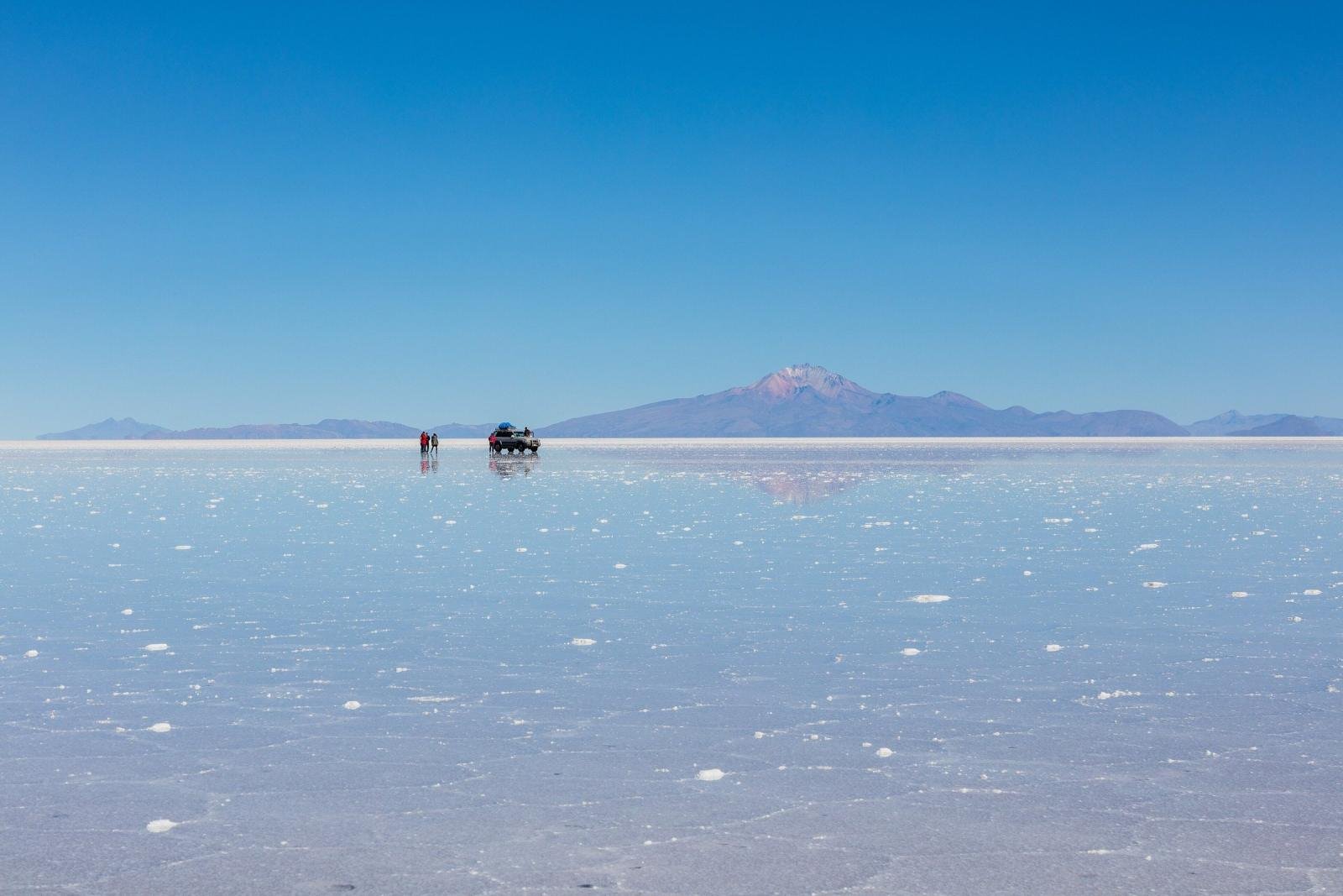 Jeep and travelers on the wet salt flat with a volcano on the horizon