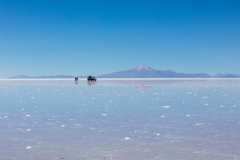 Jeep and travelers on the wet salt flat with a volcano on the horizon